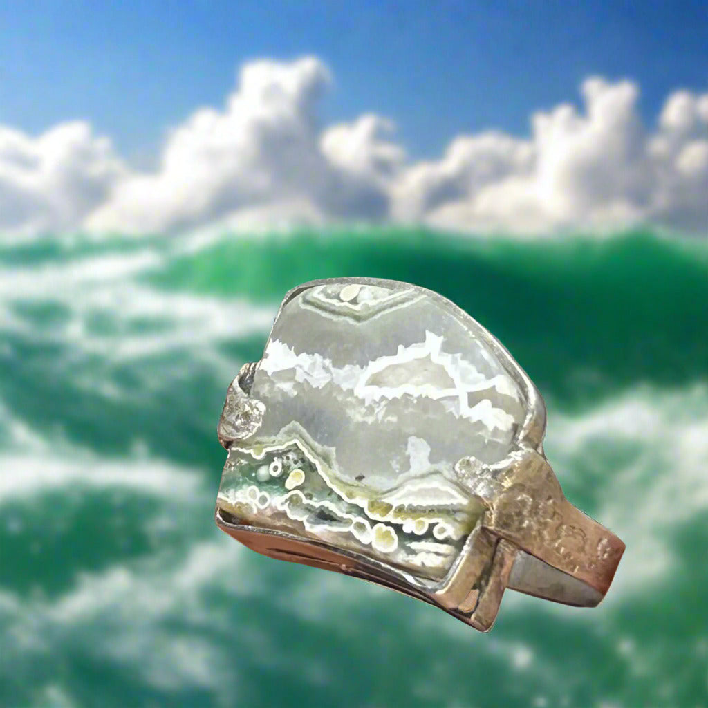 A stunning heavy rain with a slice of polished ocean Jasper, representing the sea and the sky. Textured shank continues on the pattern all the way around the ring. the stone is clear in places and has beautiful, moss green and green druzy, a sparkly quartz embedded in the stone.