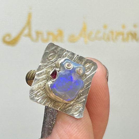 A beautiful unusual ring sporting a small Opal carving that blue in colour with two little diamonds a size and a little ruby on the right hand. The Opal is sitting on a square-ish place of silver textured to look like water